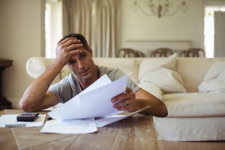 Middle-aged man sitting at home reviewing paperwork using calculator on table and pressing forehead. Professional, workspace, modern, financial, organization, comfort, introspectionの写真素材