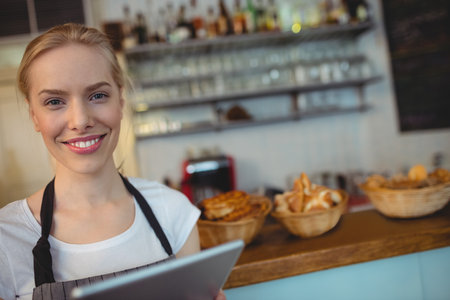 Smiling woman holding tablet behind counter in cafe arranging baskets of pastries by coffee machine. Bakery, cafe, pastry, oven, hospitality, retail, rusticの写真素材