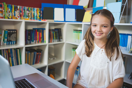 Girl wearing white blouse sitting at desk in school library using laptop near bookshelf, copy space. Educational, learning, library, study, youth, classroom, developmentの写真素材