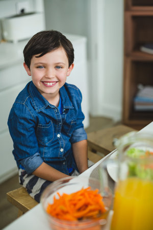 Boy smiling and sitting at dining table at home with bowl of carrots, pitcher orange juice. Child, domestic, cheerful, minimalist, wholesome, lifestyle, casualの写真素材