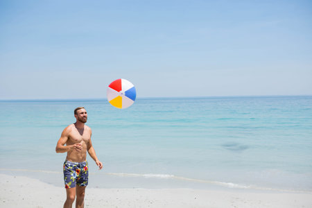 Shirtless man in floral swim trunks tossing multicolored beach ball on sand shoreline, copy space....