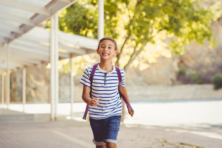 Male child running along paved walkway under white metal canopy carrying purple backpack. Joyful, energetic, outdoor, vibrant, youth, exploration, adventureの写真素材