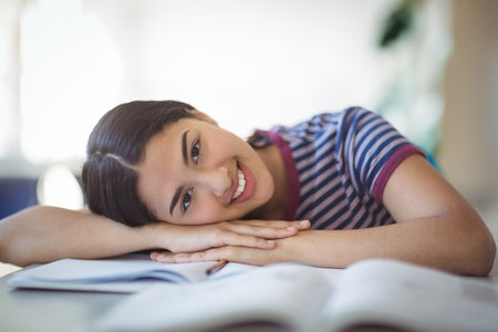 Asian teenage girl resting head on folded arms at home study desk with open textbooks. Youth, education, learning, focused, comfortable, cozy, studyingの写真素材