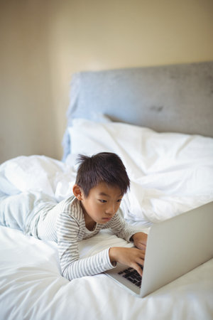 Chinese boy wearing striped pajamas lying on stomach on bed at home using silver laptop. Children, bedroom, modern, technology, comfort, relaxation, cozyの写真素材