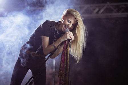 Female singer leaning into microphone stand under stage lights with smoke haze, holding scarf. Performance, musician, concert, entertainment, stage, spotlight, artistryの写真素材