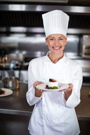 Female chef wearing coat, hat while holding dessert plate in restaurant kitchen with steel counter. Gourmet, culinary, presentation, craftsmanship, elegance, hospitality, modernの写真素材