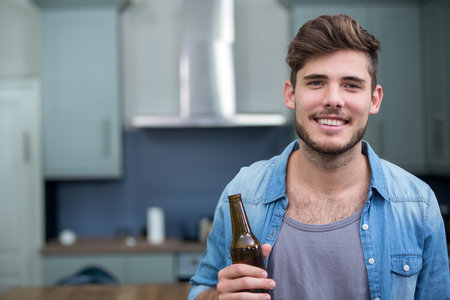 Adult man holding beer bottle and standing in home kitchen with grey cabinets, utensils, copy space. Casual, lifestyle, modern, rustic, relaxation, beverage, preparationの写真素材