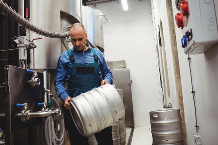 Male brewer wearing work overalls handling metal beer keg in brewery with fermentation tanks....
