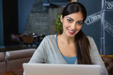 Woman smiling while typing on silver laptop at wooden cafÃ© table with chalkboard art and plant. Co-working, cafÃ©, workspace, modern, relaxed, professional, creativityの写真素材