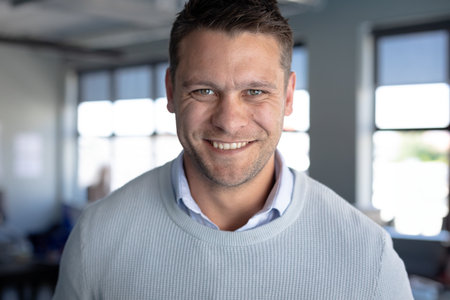 Man in his 30s wearing light-blue sweater smiling at camera in office with desks, large windows....