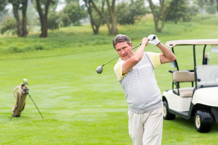Senior man swinging driver golf club on fairway in polo shirt near golf cart, copy space. Athletic, leisure, nature, outdoor, elegance, recreation, healthの写真素材