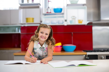 Girl writing in open notebook on kitchen countertop using pink pencil near bowls and refrigerator. Children, education, creativity, homemaking, vibrant, organized, domesticの写真素材