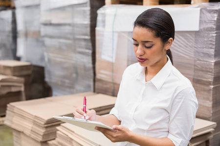 Female worker checking stock with clipboard among shrink-wrapped pallets in warehouse, copy space. Inventory, logistics, organization, industrial, professional, efficiency, bulkの写真素材