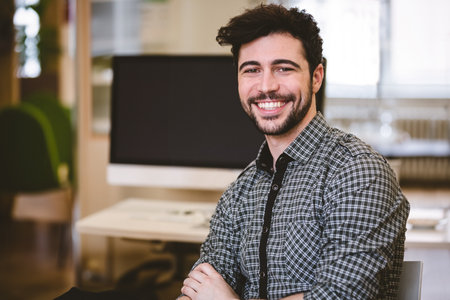 Man sitting at desk crossing arms in office workspace wearing checkered shirt with computer monitor. Professional, workspace, minimalistic, modern, business, productivity, corporateの写真素材