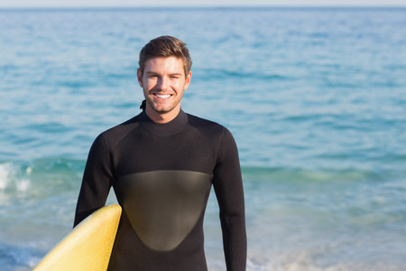 Male surfer wearing black wetsuit wading waist-deep at sandy shore holding yellow surfboard smiling. Adventure, ocean, coastline, leisure, sport, summer, vitalityの写真素材