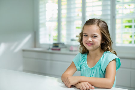 Child girl sitting at white tabletop in bright modern kitchen, smiling under window shutters. Children, family, interior, home, brightness, sunlight, minimalismの写真素材