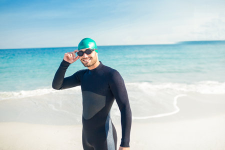 Man wearing wetsuit, swim cap lifting swim goggles on shoreline by ocean waves, copy space. Adventure, outdoor, aquatic, sport, active, fitness, coastalの写真素材