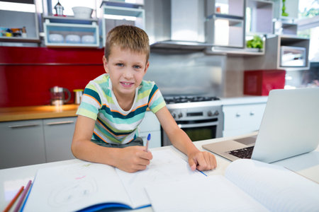 School-age boy writing in notebook with blue pen at kitchen counter with laptop, colored pencils. Children, education, creativity, workspace, domestic, lifestyle, modernの写真素材
