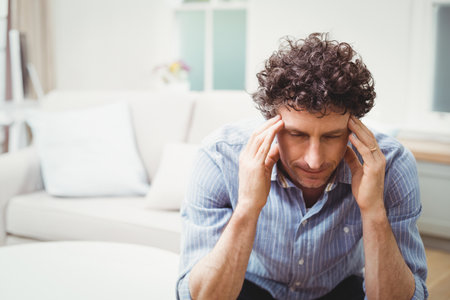 Man pressing temples while sitting on light sofa in living room with flower vase, copy space. Conflict, contemplation, comfort, relaxation, interior, muted, serenityの写真素材