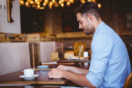 Man sitting at wooden table in coffee shop wearing striped shirt holding white cup, copy space. Cafe, relaxation, cozy, warmth, rustic, comfort, leisureの写真素材