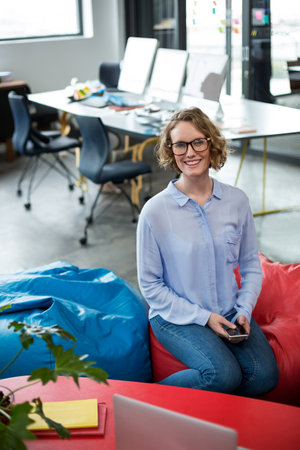 White woman in twenties using smartphone while sitting on beanbag chairs in open office with laptop....