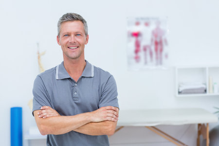 Mature male therapist standing arms crossed in therapy room next to treatment table. Professional, healthcare, wellness, rehabilitation, anatomy, therapy, medicalの写真素材