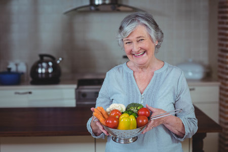 Senior woman smiling while holding metal colander of fresh vegetables in home kitchen. Health, nutrition, harvest, freshness, culinary, domestic, vibrantの写真素材