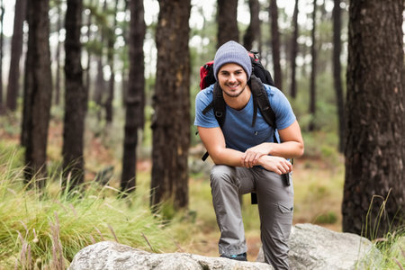 Man kneeling on large boulder in pine forest, wearing beanie and hiking boots carrying red backpack....