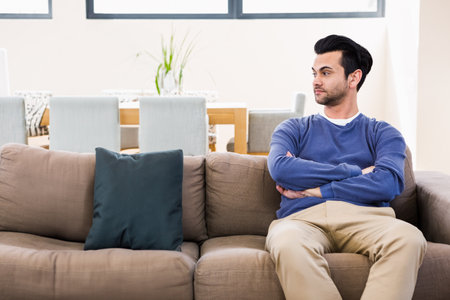 Man sitting on sofa folding arms beside teal pillow at home near dining table, copy space. Modern, relaxation, cozy, interior, minimalism, comfort, homeの写真素材
