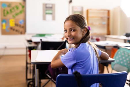 Girl child sitting at school desk turning around smiling, looking at camera with notebook, pencil. Children, education, learning, classroom, academic, student life, youthの写真素材