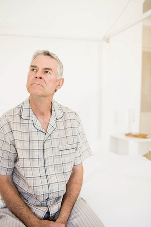 Senior man wearing plaid pajamas sitting on bed in bedroom beside table with lamp, copy space. Comfort, relaxation, home, tranquility, minimalism, wellness, lifestyleの写真素材