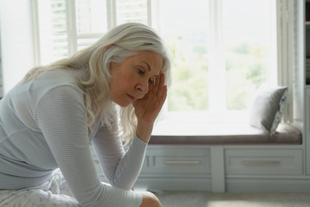 Senior woman sitting on bench under window shutters in bedroom, pressing hand to temple, copy space. Retirement, relaxation, comfort, cozy, mindfulness, sanctuary, tranquilityの写真素材