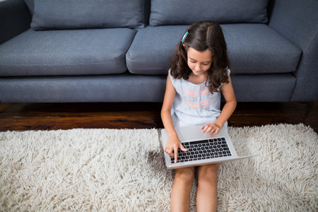 Girl child sitting on white rug in front of dark grey sofa working on silver laptop. Playful, cozy,...