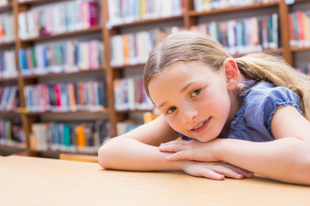 Female child leaning on wooden table gazing at camera in library area with bookshelves, copy space. Child, library, reading, education, exploration, study, learningの写真素材