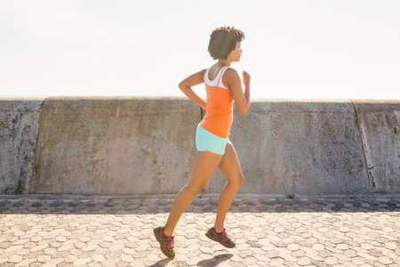 Runner jogging along hexagonal paver walkway beside concrete seawall under bright sunlight. Athletic, wellness, outdoor, vibrant, summer, fitness, landscapeの写真素材