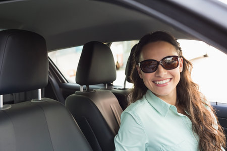 Smiling female wearing sunglasses and holding steering wheel, leaning toward window in parked car....
