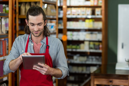 Male clerk in red apron tapping tablet by wooden shelves with boxes, bottles, countertop and fridge....