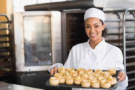 African American woman pastry chef holding tray of golden pastries at bakery prep table, copy space. Pastry, bakery, culinary, industrial, equipment, chef, confectionsの写真素材