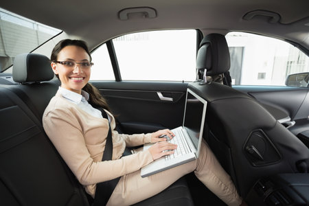 Woman sitting in backseat of modern sedan wearing eyeglasses and seatbelt typing on laptop on lap. Luxury, mobility, technology, business, commute, productivity, professionalの写真素材