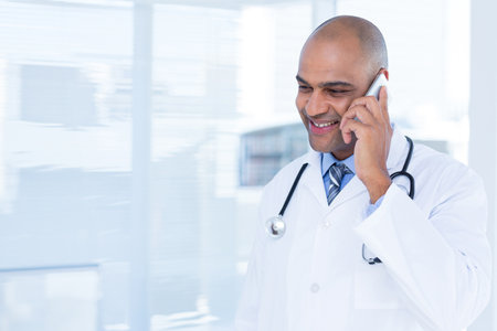 Doctor wearing lab coat and stethoscope is speaking on smartphone near urban skyline window blinds. Medical, healthcare, business, professional, modern, urban, communicationの写真素材