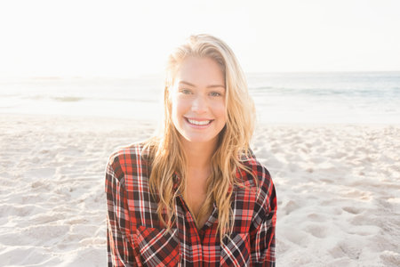 Woman smiling standing on sandy beach near water's edge wearing plaid shirt under backlit sunlight. Outdoors, leisure, fashion, lifestyle, nature, relaxation, vibrantの写真素材