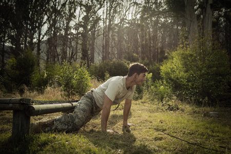 soldier doing incline push-up on grass with feet on wooden beam forest clearing, sunlight filtering. Military, training, fitness, outdoor, obstacle, endurance, strengthの写真素材