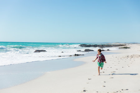 Boy in plaid shirt, green shorts running barefoot on beach leaving footprints by rocks, copy space. Harbor, leisure, adventure, nature, outdoor, vibrant, youthfulの写真素材