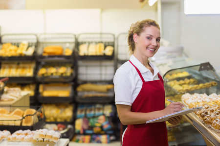 Female bakery worker holding clipboard inspecting bread racks and pastries at counter, copy space. Pastries, bread, retail, artisan, dairy-free, rustic, seasonalの写真素材