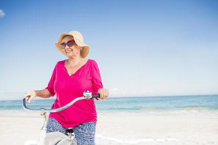Senior woman standing on beach wearing straw sun hat and sunglasses holding white cruiser bicycle. Vanity, leisure, relaxation, outdoor, tranquility, wellness, vibrantの写真素材
