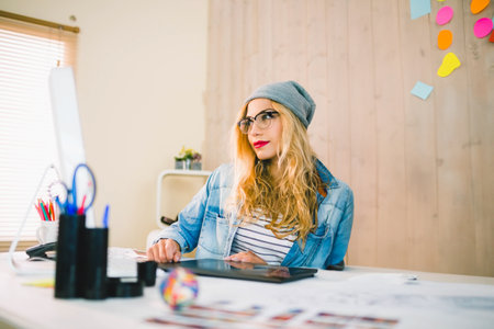 Graphic designer wearing glasses and beanie sketching color art on tablet at studio desk. Creative workspace, artistic tools, modern interior, freelance, vibrant, innovative, productivityの写真素材