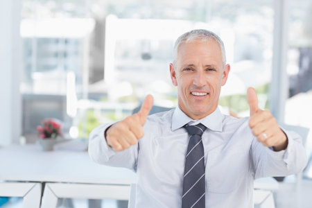 Man in business attire giving thumbs-up at office desk with potted plant showing red flowers.の写真素材