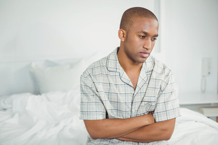 African American man sitting on bed in bedroom under intercom in plaid pajamas, copy space. Relaxation, comfort, tranquility, bedroom, lifestyle, minimalism, wellnessの写真素材