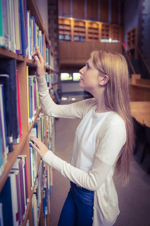 Female student is browsing wooden bookshelves and reaching for book in library hall under warm glow. Literature, education, research, interior, renovation, focus, inspirationの写真素材