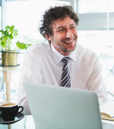 Mature man in business attire working on laptop at workspace by window with coffee cup, notebook. Professional, urban, natural, modern, relaxed, productivity, brightの写真素材